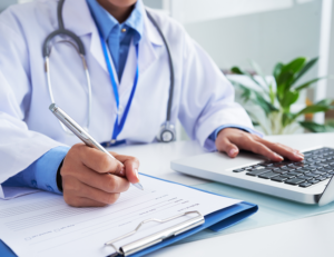 Médico com jaleco e estetoscópio anotando em caderno e digitando em notebook em cima de uma mesa branca com um vaso de flores desfocado ao fundo.
